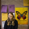30 Under 30 honoree Michelle Ferguson Shore smiling and sitting in front of a wall covered in brown craft paper, featuring a butterfly poster and yellow and purple paper